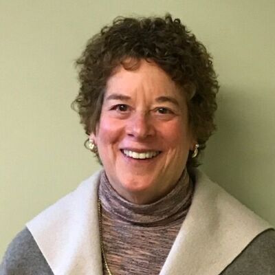 A smiling woman with curly hair wearing a gray jacket over a turtleneck sweater stands in front of a plain light green background, embodying the confidence and poise often seen in members of the leadership team. Saint Joseph's College of Maine