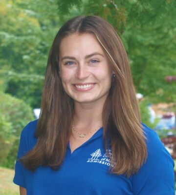 A person with long brown hair smiles at the camera. They are wearing a blue polo shirt with a college logo. The background is a blurred outdoor setting with greenery. Saint Joseph's College of Maine