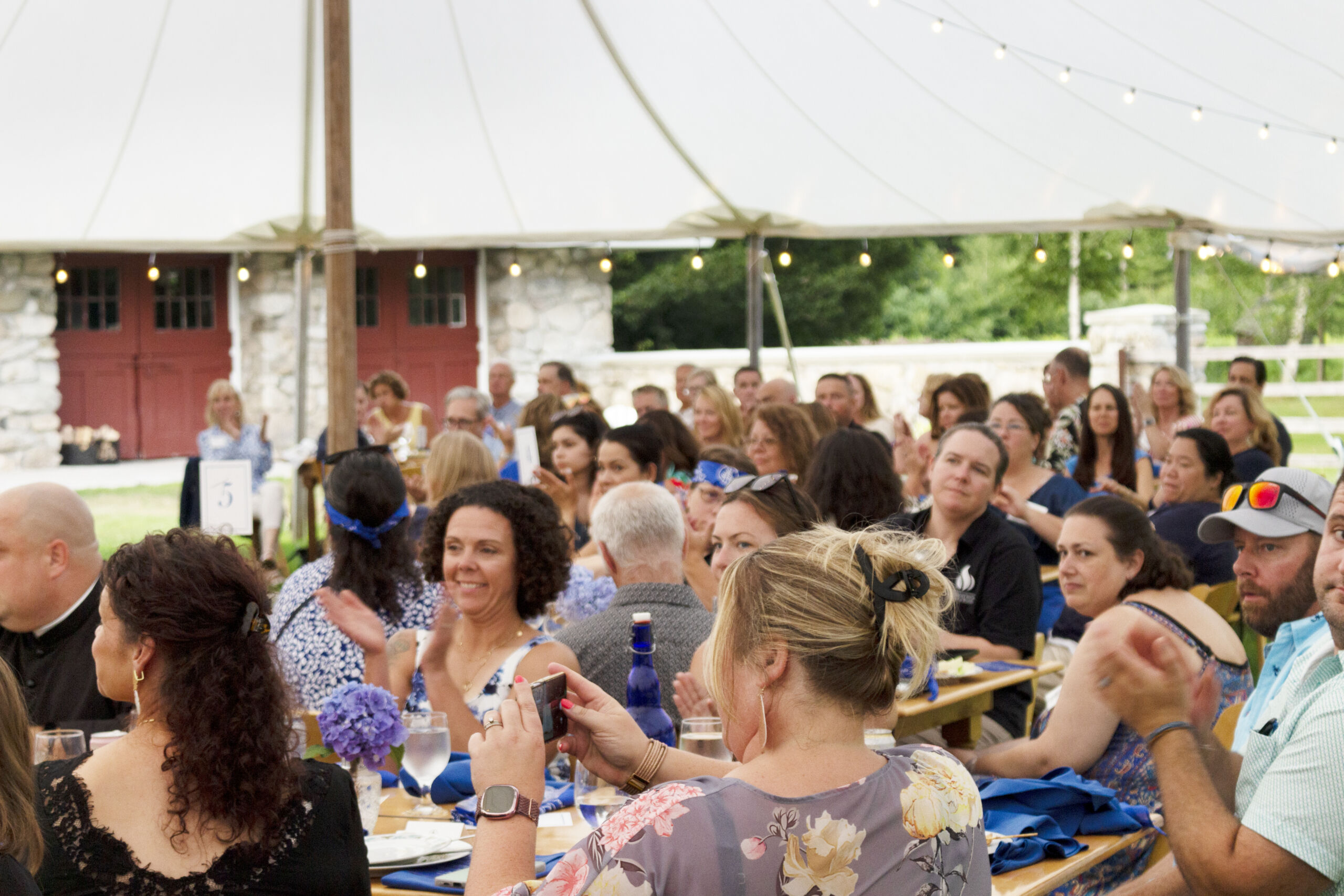 A large group of people seated at tables under a tent, engaging in conversation and clapping. String lights and a stone building are in the background. Saint Joseph's College of Maine