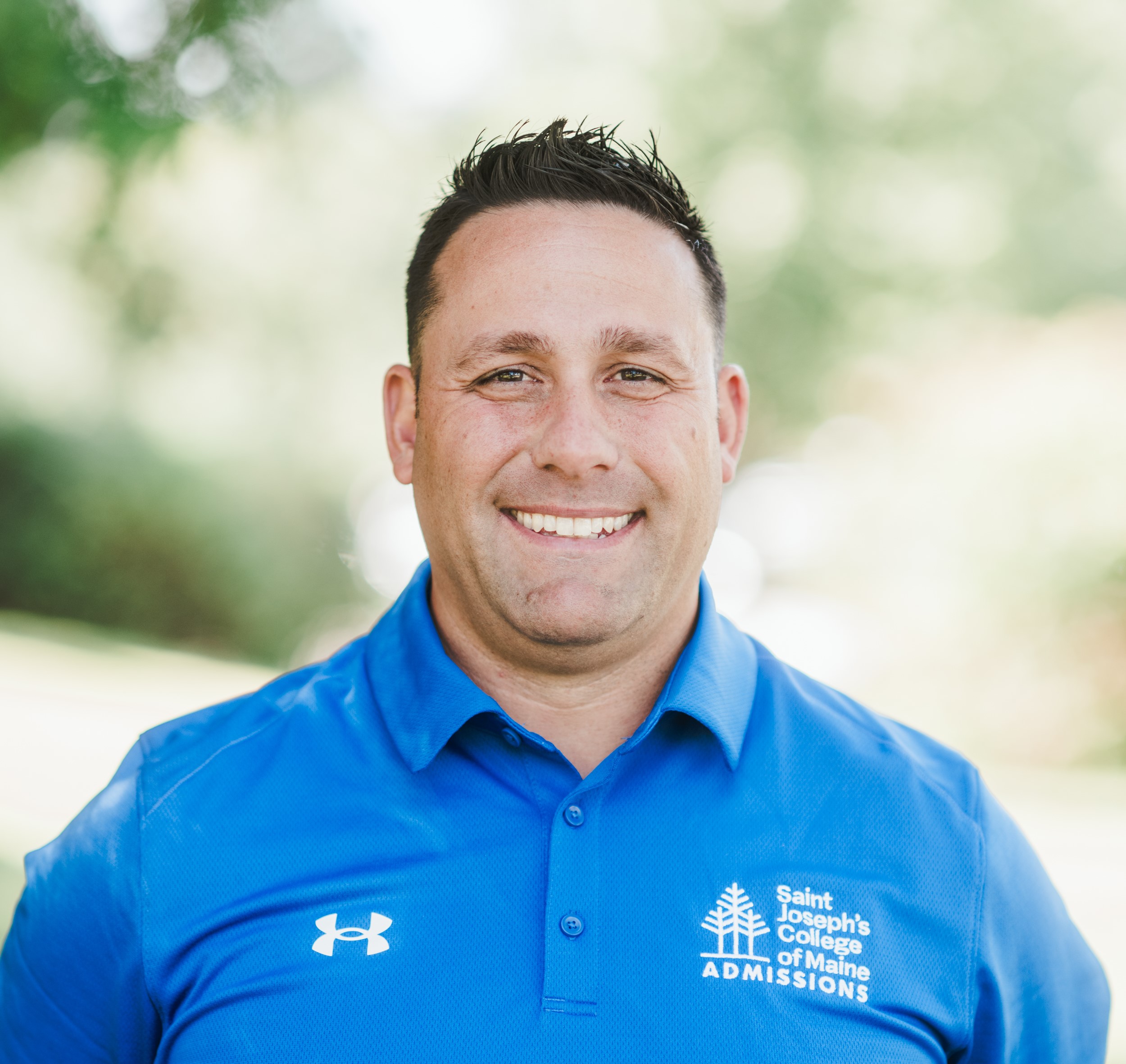 A person wearing a blue polo shirt with a Saint Joseph's College of Maine Admissions logo smiles outdoors. Saint Joseph's College of Maine