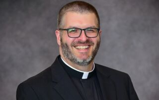 Reverend Patrick Finn, a chaplain at Saint Joseph’s College of Maine, sports short hair, glasses, and a beard. He wears a black clerical collar and suit while smiling against a grey background. Saint Joseph's College of Maine