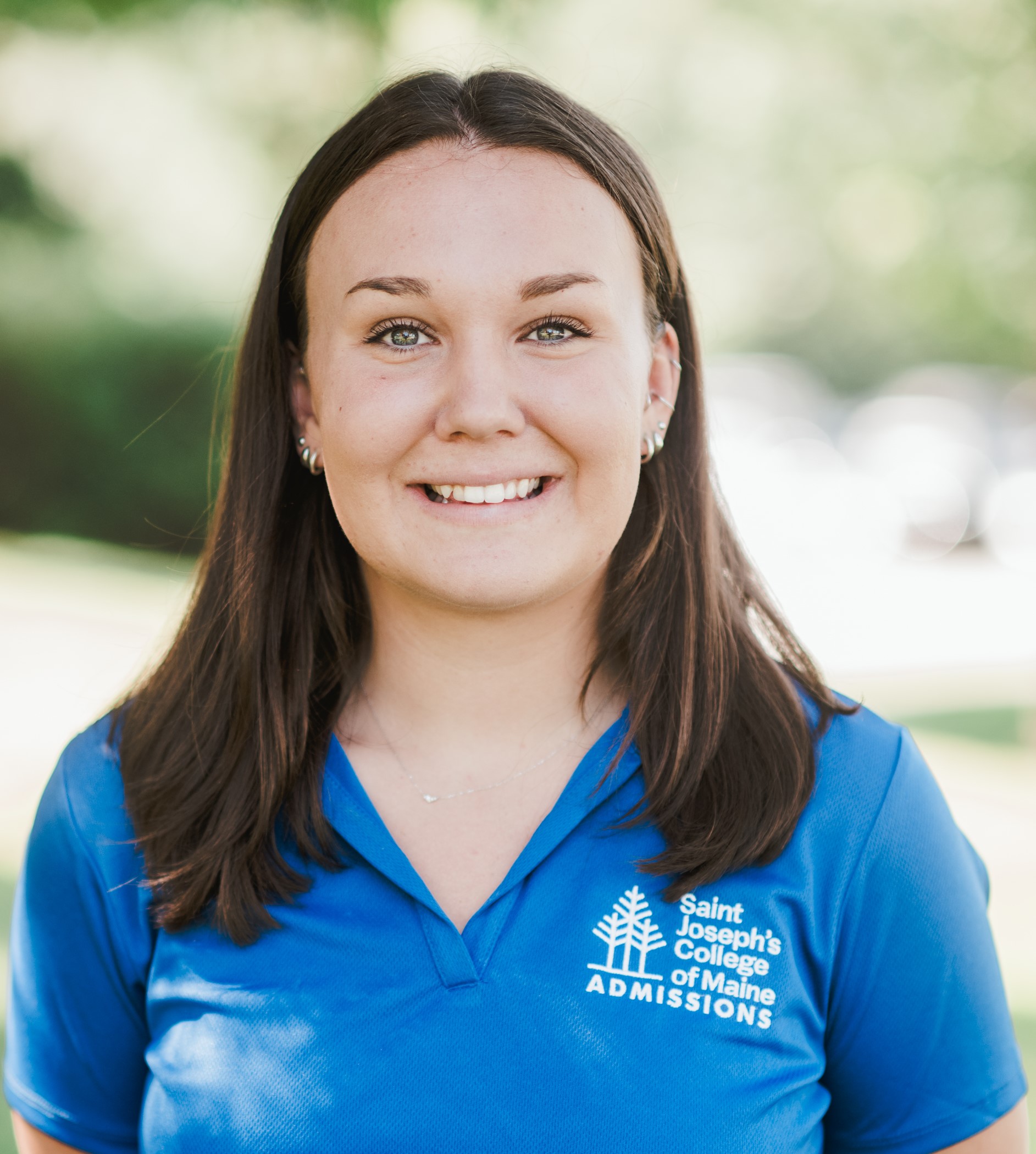 A person with long brown hair wearing a blue "Saint Joseph's College of Maine Admissions" shirt smiles outdoors. Saint Joseph's College of Maine