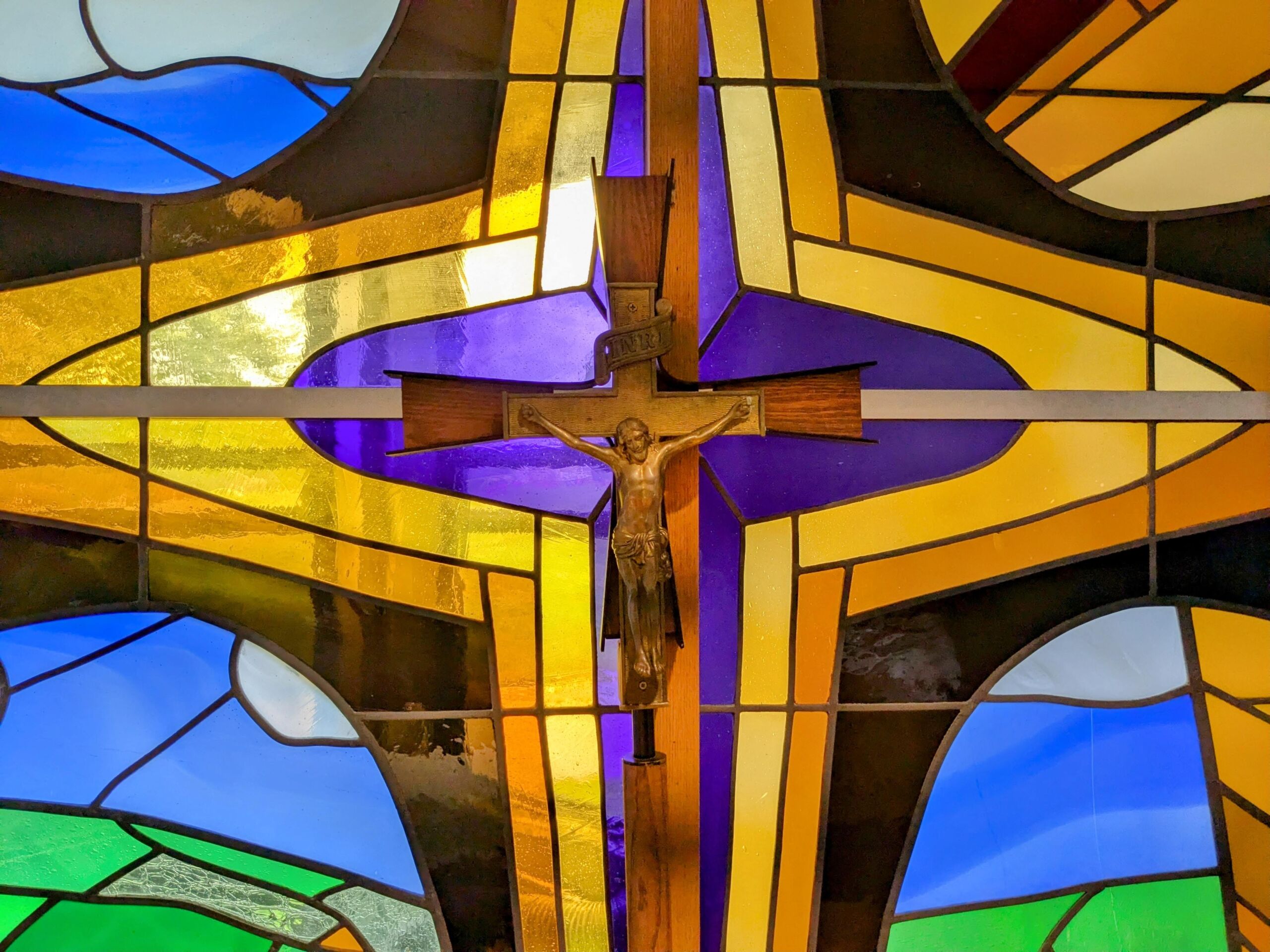 A crucifix is centered within a colorful stained glass window featuring geometric patterns and a mix of blue, green, yellow, and purple sections that reflect the diverse spirit of Life and Community. Saint Joseph's College of Maine