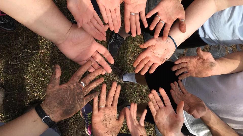 A group of community members stands in a circle, each extending their hands towards the center. Some hands are clean, while others are dirty, symbolizing the diverse experiences and stories that make up our shared life and community. Saint Joseph's College of Maine