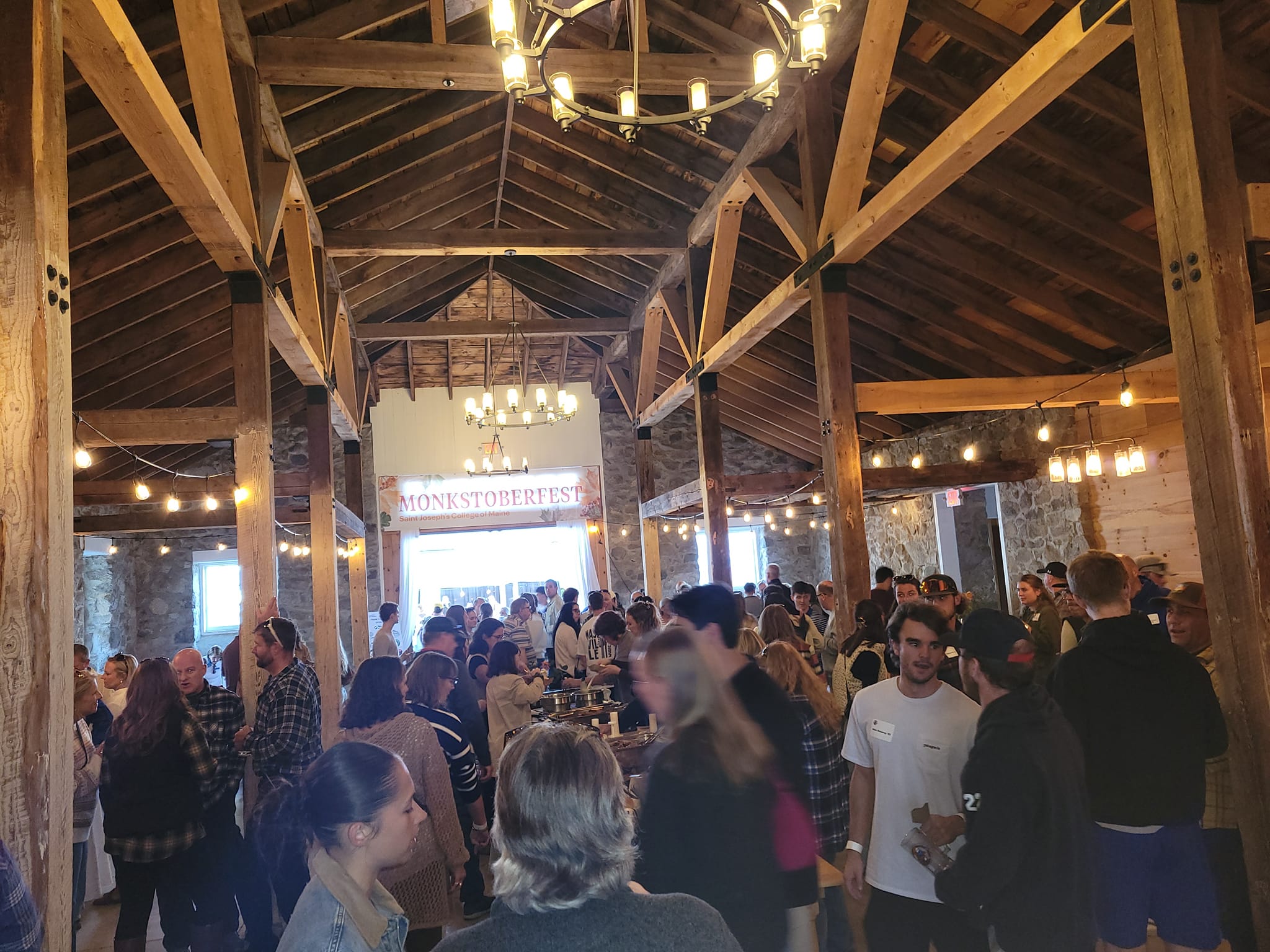 A large crowd gathers inside a rustic, wooden-beamed hall for an event called Monkstorefest. Tables are set up with people mingling and chatting. Saint Joseph's College of Maine