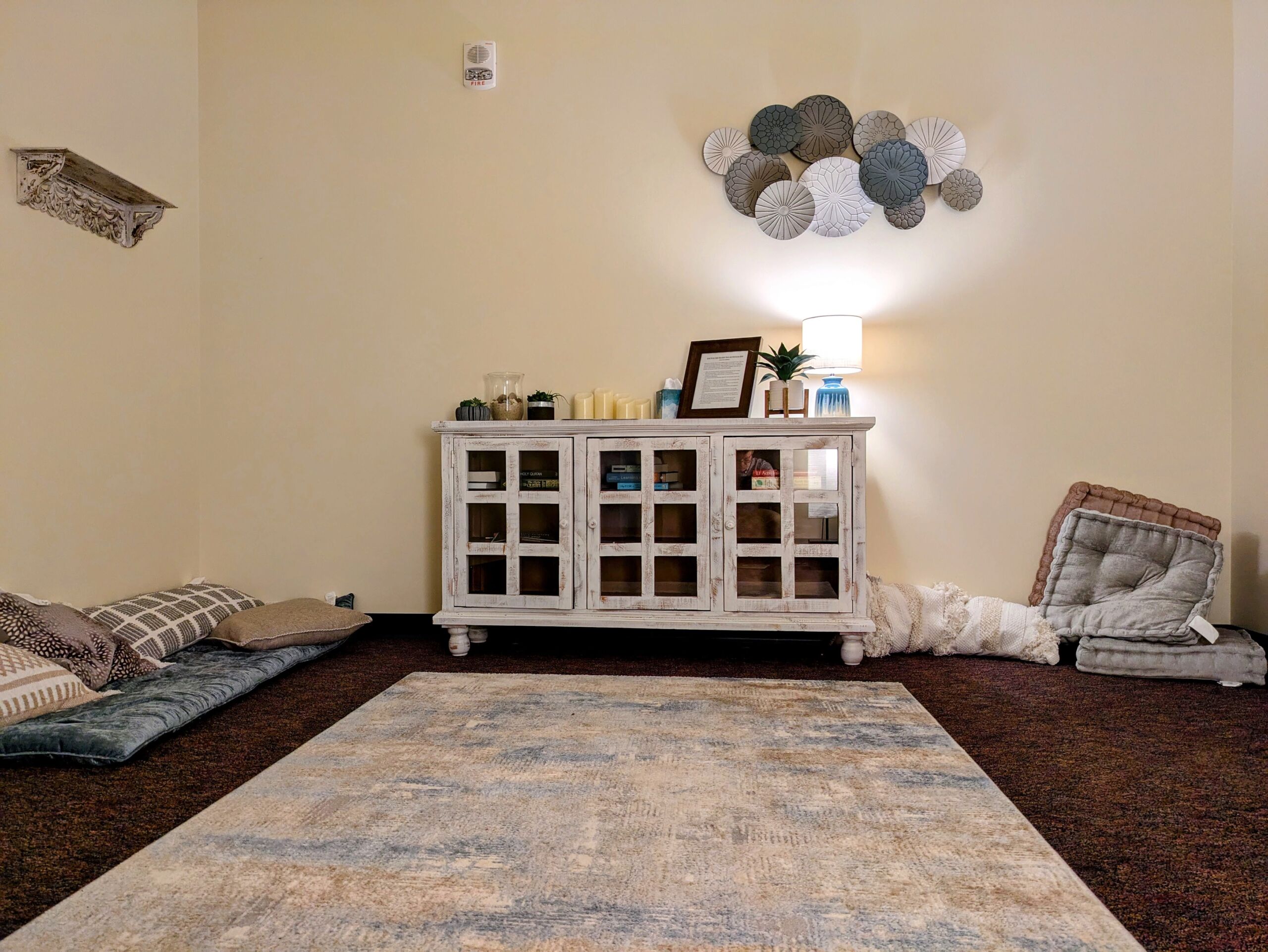 A quiet room with a white cabinet, a lamp, decorative wall art, cushions on the floor, and a beige rug on brown carpet—perfect for community members to gather and enjoy moments of peaceful Campus Life. Saint Joseph's College of Maine