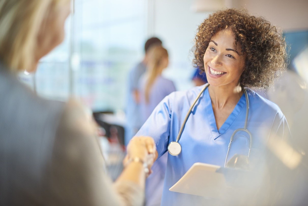 A medical professional in blue scrubs and a stethoscope smiles and shakes hands with a person, while holding a clipboard. Saint Joseph's College of Maine