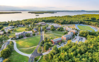 Aerial view of Saint Joseph’s College campus in Maine, showcasing various buildings and sports facilities surrounded by lush greenery, adjacent to a large body of water during daytime. Saint Joseph's College of Maine