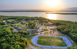 Aerial view of a campus with buildings, a running track, and a sports field for aspiring soccer players and Soccer clinics, surrounded by trees near a large body of water at sunset. Saint Joseph's College of Maine