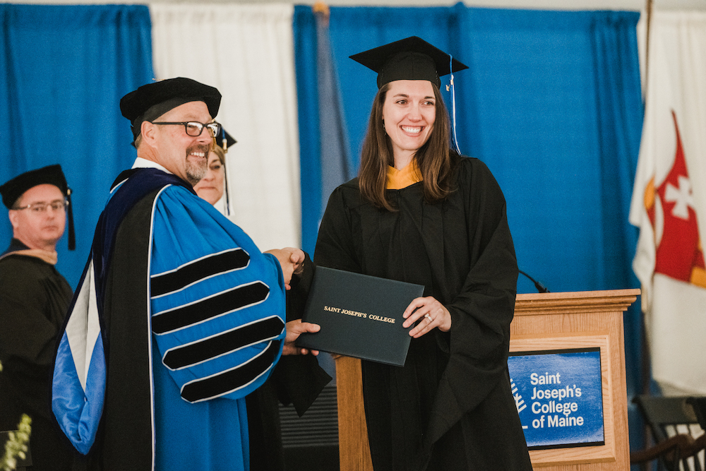 A graduate in a black cap and gown from the Class of 2024 receives a diploma from a faculty member at an SJC graduation ceremony. Saint Joseph's College of Maine