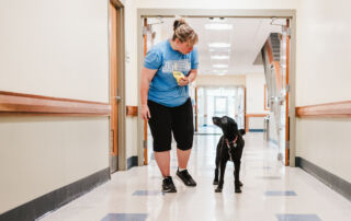 A woman wearing a blue shirt and black shorts walks down a hallway with a black dog, holding a yellow object in her hand. Saint Joseph's College of Maine