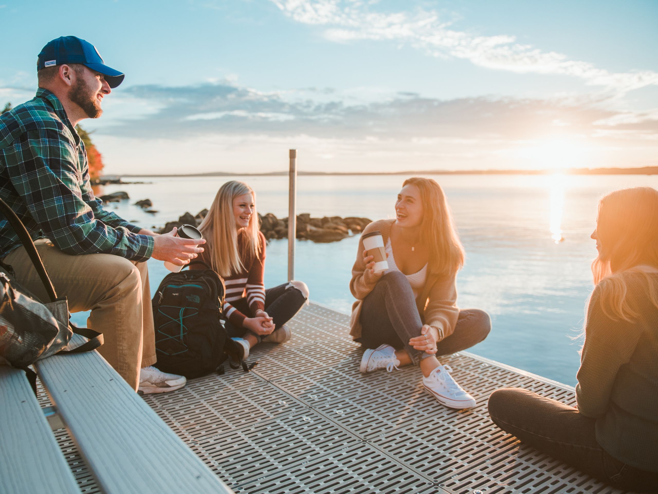 Four people sit on a dock by a lake at sunset, talking and laughing. Saint Joseph's College of Maine