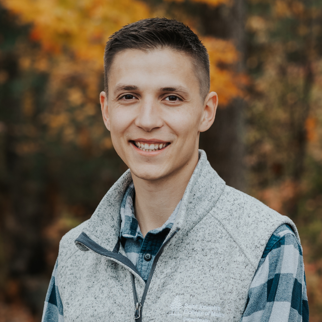 A person wearing a gray vest and plaid shirt smiles outdoors with a background of autumn leaves. Saint Joseph's College of Maine