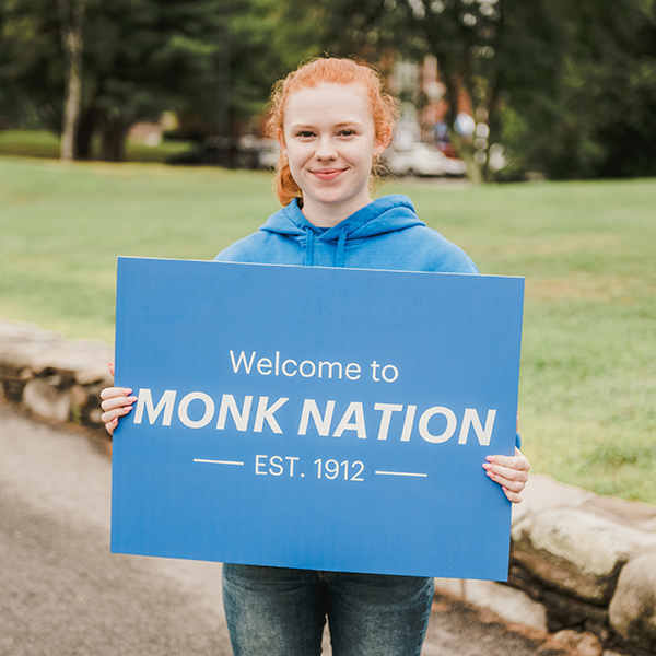Student holds a sign for new students that says: Welcome to Monk Nation
