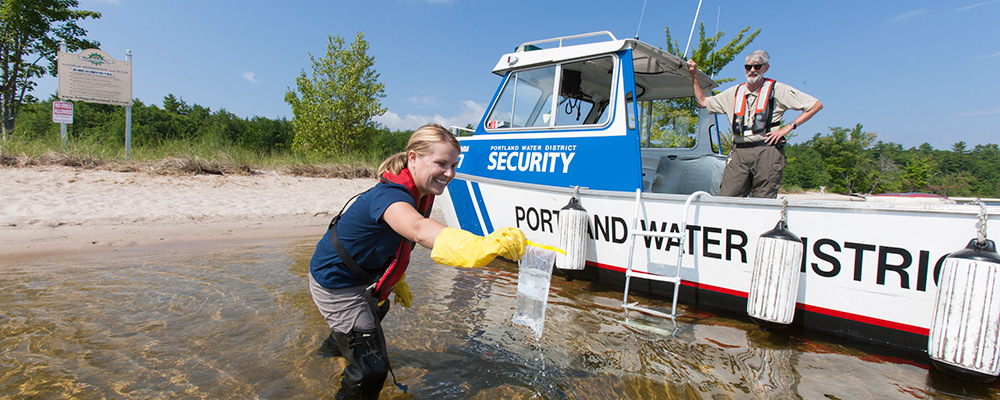 Woman taking a water sample with the Portland Water District boat in the background